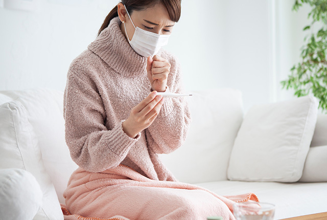 ソファに座り体温計を見て咳をするマスク姿の女性